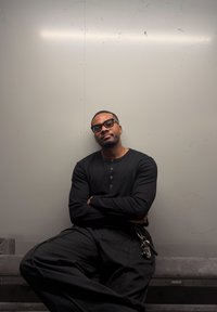 Chemise noire à manches longues côtelée, pantalon noir ample et lunettes ; assis contre un mur lisse gris clair, avec un porte-clés visible.