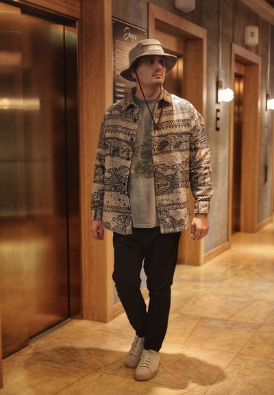 Camisa beige con estampado y acentos oscuros, camiseta gris, pantalones negros y zapatillas blancas; lleva un sombrero beige. El fondo incluye elementos de madera y metálicos.