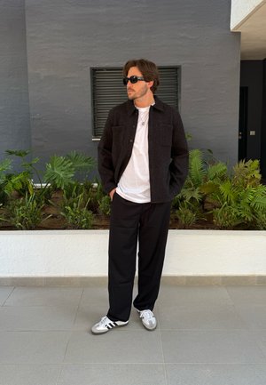Chaqueta negra de botones, camiseta blanca y pantalones anchos negros. Zapatillas grises con rayas negras. Entorno interior con plantas.