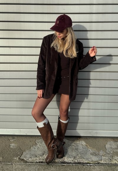 Blazer de pana marrón sobre un vestido negro ajustado, combinado con botas marrones hasta la rodilla y calcetines con acentos blancos, sobre un fondo gris con rayas.