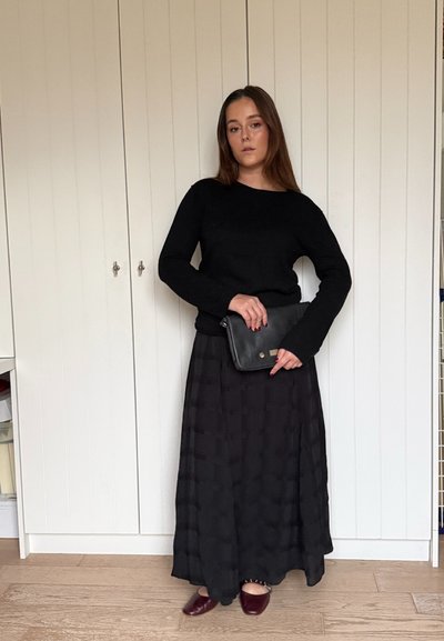 Suéter de punto negro, falda larga negra plisada con patrón de cuadrícula, bolso negro y zapatos burdeos. Sobre un suelo de madera clara y frente a armarios blancos.