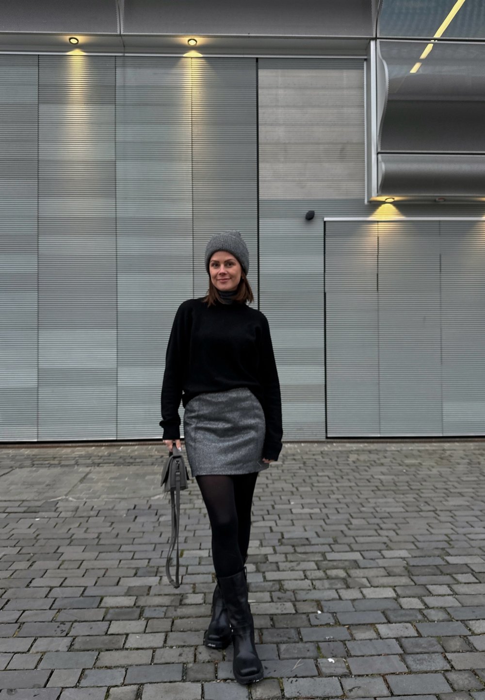 Μαύρο πουλόβερ με ψηλό λαιμό, ασημί μίνι φούστα, μαύρες καλσόν και χοντρές μαύρες μπότες. Γκρι σκουφί και μικρή γκρι τσάντα. Φόντο από κυβόλιθους.