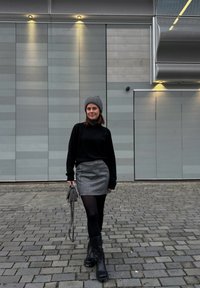 Μαύρο πουλόβερ με ψηλό λαιμό, ασημί μίνι φούστα, μαύρες καλσόν και χοντρές μαύρες μπότες. Γκρι σκουφί και μικρή γκρι τσάντα. Φόντο από κυβόλιθους.