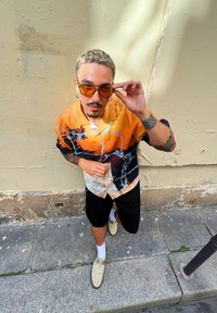 Κοντομάνικο πουκάμισο με gradient μοτίβο πορτοκαλί προς μαύρο, με ελαφριά μπλε λουλουδάτα �αξεσουάρ. Φοριούνται μαύρες βερμούδες και ανοιχτόχρωμα παπούτσια.