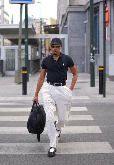 Camisa polo negra, pantalones blancos, zapatos negros con acentos blancos. Lleva un bolso de lona negro y camina por un paso de peatones en un entorno urbano.