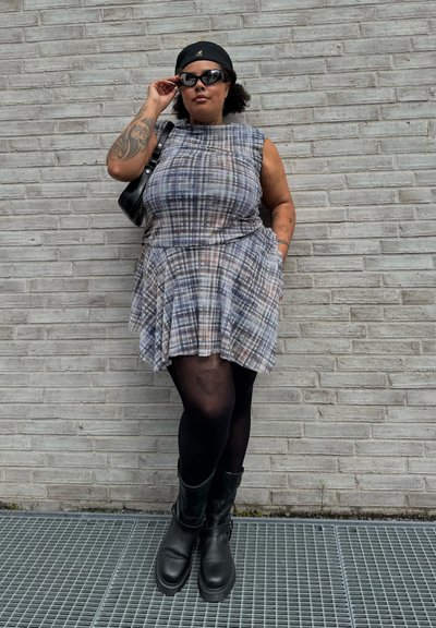 Vestido de cuadros en gris, rosa y azul, con falda acampanada y diseño sin mangas. Medias negras y botas negras de suela gruesa. Bolso negro y gafas de sol.