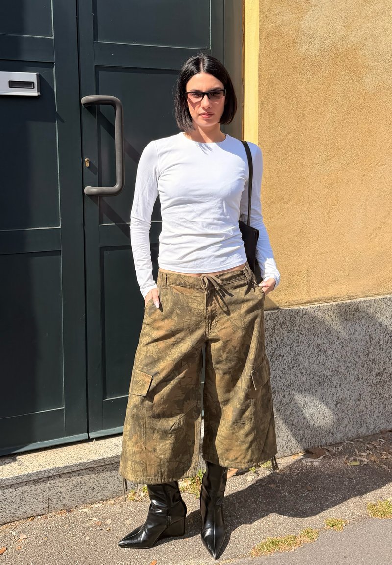 Lühikeste mustade juustega naine, kandes päikeseprille, valget pikade varrukatega särki, laiu camo-pükse ja musti teravate säärega saapaid, seisab tumeda ukse ja kollase seina ääres.