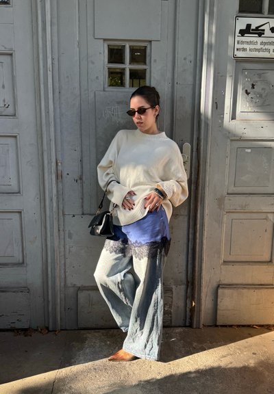 Suéter crema de gran tamaño, superpuesto a una blusa de satén azul con borde de encaje negro, combinado con jeans de pierna ancha y zapatos marrones de punta.
