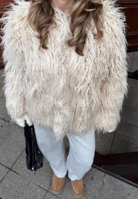Veste en fausse fourrure crème avec une texture longue imitant les poils. Associée à un pantalon gris clair et des chaussures marron clair, tenant un sac noir.