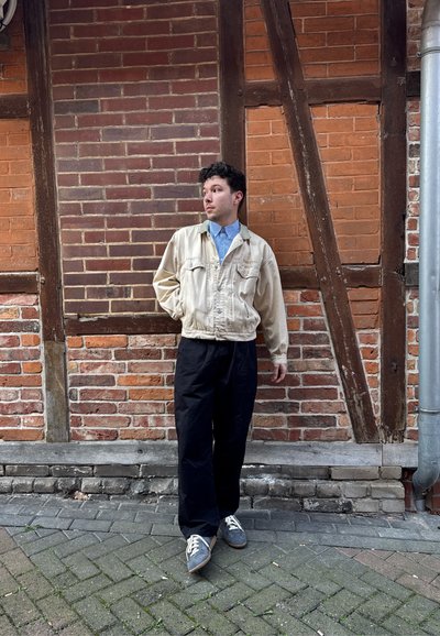 Joven con chaqueta beige y pantalones negros está de pie mirando hacia la izquierda frente a una pared de ladrillo y madera al aire libre.