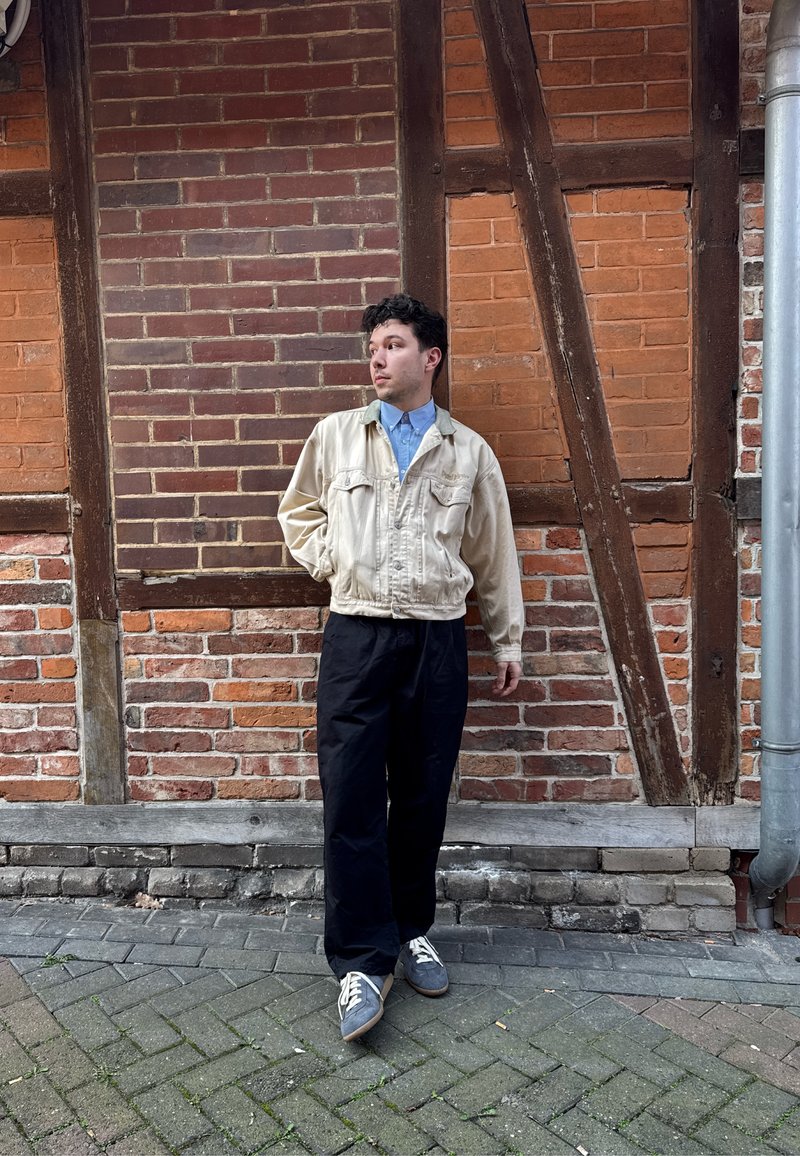 Jeune homme en veste beige et pantalon noir se tenant debout et regardant à gauche devant un mur en briques et en bois à l'extérieur.
