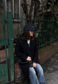 Γυναίκα με μαύρο παλτό και καπέλο, κάθεται σε τοίχο από τούβλα δίπλα σε πράσινο μεταλλικό φράχτη και ξυλεμένο δέντρο, κοιτάζει προς τα δεξιά.