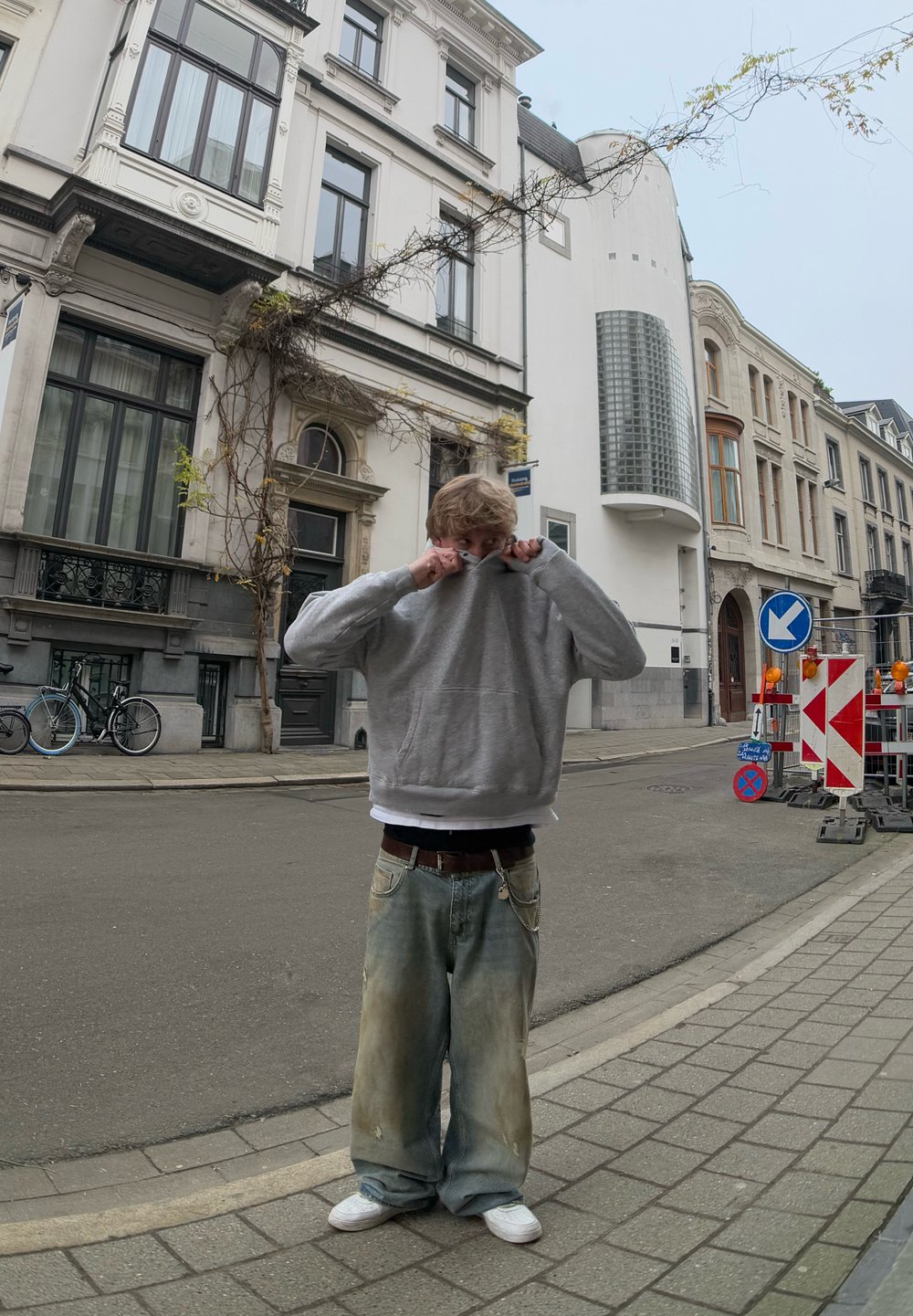 Γκρίζα κουκούλα φούτερ, χαλαρά ανοιχτό μπλε τζιν με λεκέδες, λευκά sneakers, στέκεται σε έναν δρόμο με ιστορικά κτίρια και ποδήλατα.