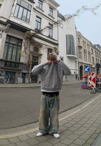 Γκρίζα κουκούλα φούτερ, χαλαρά ανοιχτό μπλε τζιν με λεκέδες, λευκά sneakers, στέκεται σε έναν δρόμο με ιστορικά κτίρια και ποδήλατα.