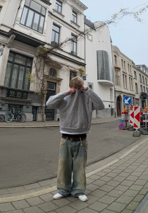 Siva kapucca s kapuco, ohlapne svetlo modre kavbojke z madeži, bele teniske, stoji na ulici s zgodovinskimi stavbami in kolesi.