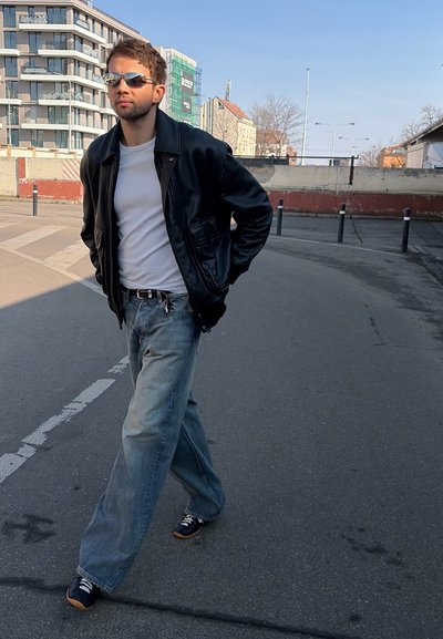 Joven con gafas de sol, chaqueta de cuero negra, camisa blanca y vaqueros holgados caminando por una calle de la ciudad con edificios modernos al fondo.
