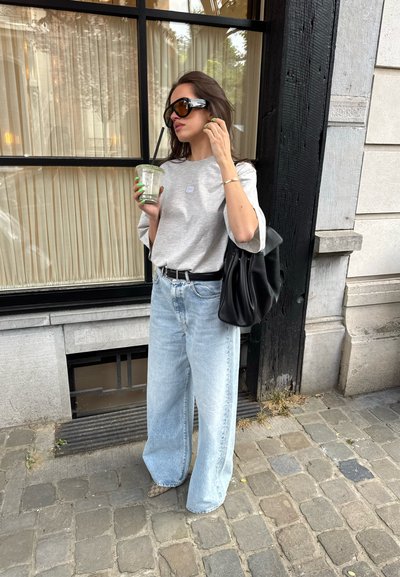 Camiseta gris de gran tamaño con un pequeño logo, jeans de pierna ancha de cintura alta en azul claro, bolso negro y gafas de sol. Sosteniendo una bebida verde.