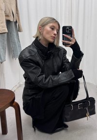 Femme blonde accroupie portant une veste en cuir noire et un pantalon, tenant un sac à main noir avec des clés, prenant un selfie dans un miroir avec un smartphone.