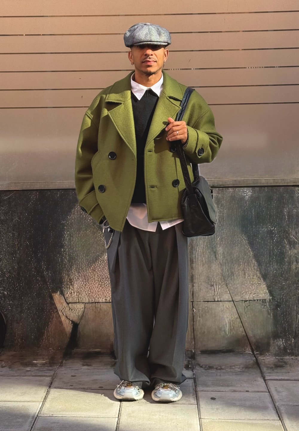 Groene oversized dubbeldoorlopende jas, zwarte trui, witte blouse, losse grijze broek, grijze pet, zwarte tas en gestructureerde sneakers.