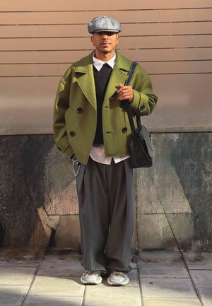 Grüner, oversized Doppelkamera-Mantel, schwarzer Pullover, weißes Hemd, lockere graue Hose, graue Kappe, schwarze Tasche und gemusterte Sneakers.