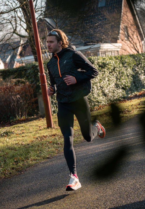 Hombre con ropa deportiva negra y zapatillas blancas corriendo por un camino pavimentado con arbustos y casas al fondo.