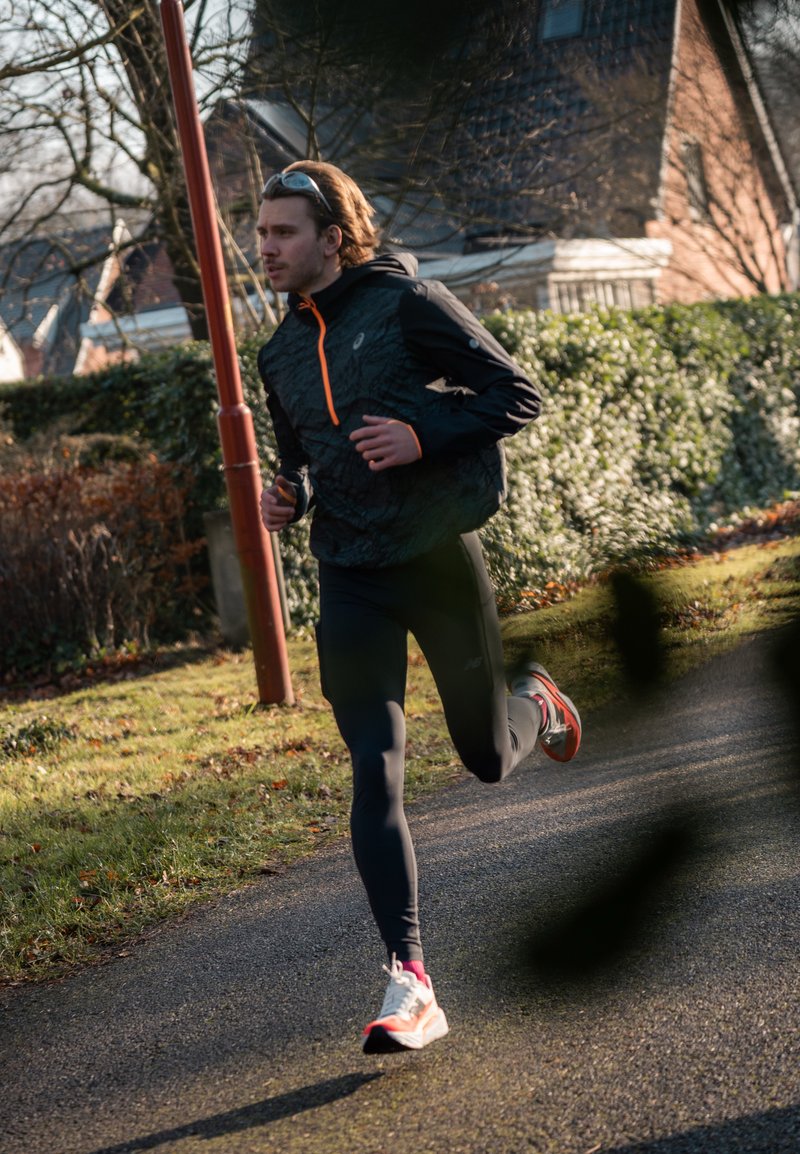 Vyras juodo bėgimo aprangos ir baltų sportbačių bėgioja pavedintu taku, fone matosi krūmai ir namai.