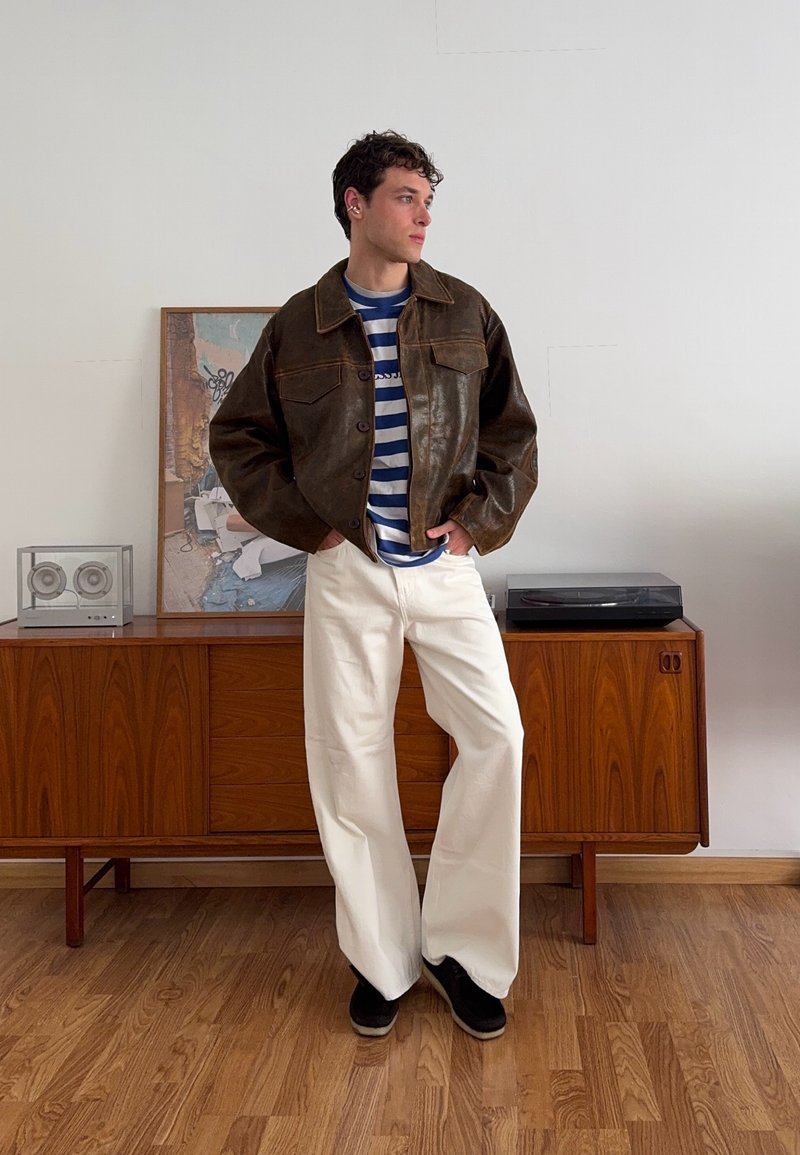 Brown leather jacket with a large collar, striped blue and white shirt, and white wide-leg trousers. The setting includes wooden furniture and a vinyl record player.
