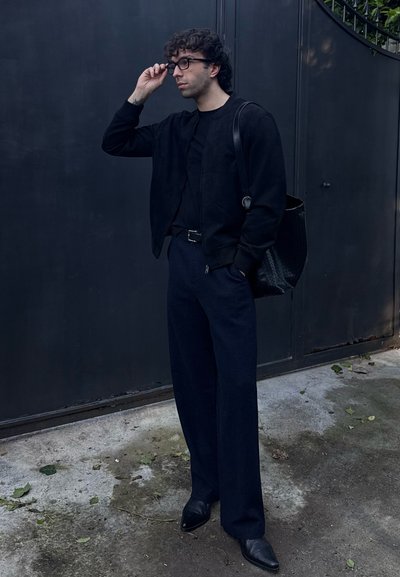 Chaqueta bomber negra sobre una camisa negra, combinada con pantalones anchos oscuros y zapatos de tacón negros. Accesorios con un bolso negro texturizado.