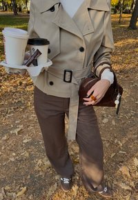 Béžový trenčkot s černými knoflíky a páskem, hnědé kalhoty s širokými nohavicemi, drží kávové kelímky v držáku, hnědá kabelka s detailním střapcem.