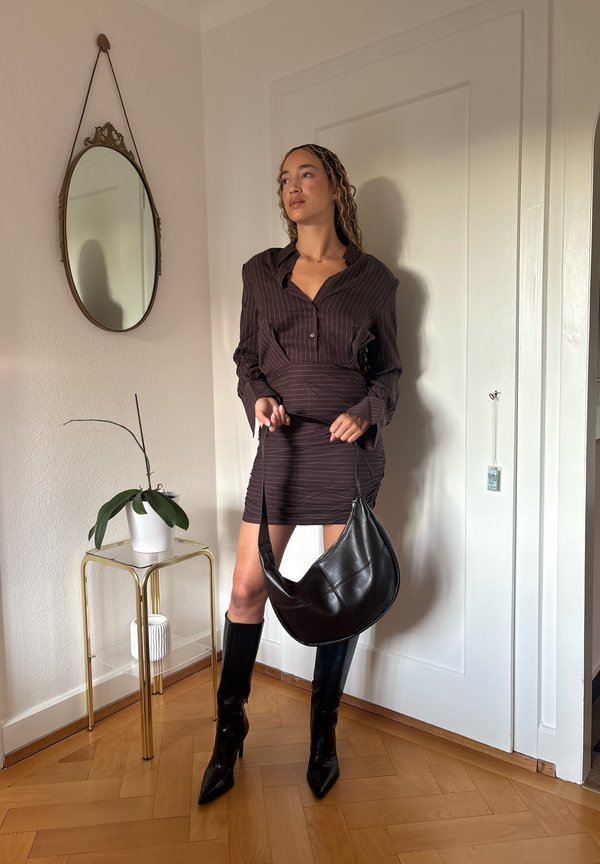 Femme en robe sombre à fines rayures et bottes noires tenant un sac en cuir noir, debout près d'un miroir et d'une petite table avec une plante.