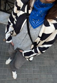 Personne portant une veste à motifs noir et blanc, une écharpe bleue à motifs autour du cou, un cardigan gris boutonné, un pantalon sombre et des baskets blanches sur un trottoir carrelé.
