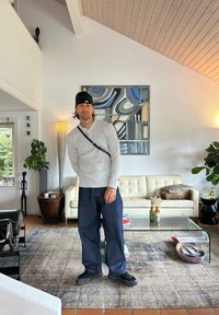 Uomo con berretto nero, maglione grigio e pantaloni larghi scuri, in piedi in un moderno soggiorno con divano beige, tavolo in vetro, piante e arte murale astratta.