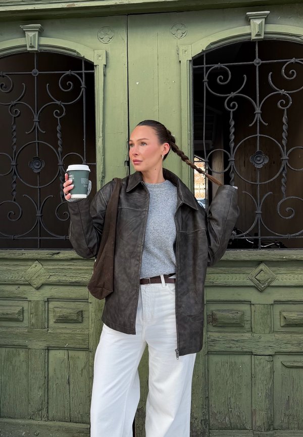 Femme en pull gris et veste en cuir foncé tenant une tasse de café et jouant avec sa tresse devant une porte en bois vert décorative.