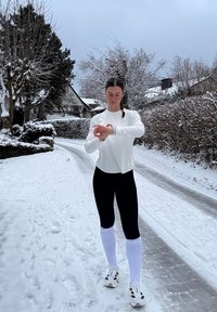 Haut blanc à manches longues, leggings noirs et chaussettes blanches montant jusqu'aux genoux. Détails réfléchissants et chaussures de course. Sentier enneigé et arbres en arrière-plan.