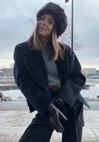 Młoda kobieta w zimowych ubraniach: ciemnym płaszczu, futrzanej czapce, rękawiczkach i szarym swetrze stoi na zewnątrz nad brzegiem wody w pochmurną pogodę.