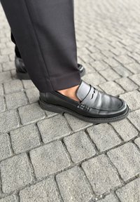 Zwarte leren loafers met elastische zijaccenten, vlakke zolen en een gladde afwerking, gedragen met donkere broeken op een kinderkopjesoppervlak.