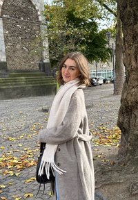 Abrigo gris de lana con botones, cinturón atado y bufanda blanca con flecos. Bolso oscuro y pantalones vaqueros azules. Camino empedrado con hojas amarillas.