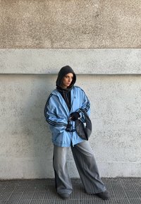 Femme portant un sweat à capuche noir sous une veste Adidas bleu clair, un pantalon gris à jambes larges, des gants noirs, tenant un sac en mesh noir, appuyée contre un mur.
