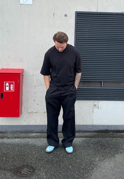 Camiseta negra de gran tamaño, pantalones negros holgados y zapatos de color azul claro. De pie contra una pared gris cerca de una caja de seguridad contra incendios roja.