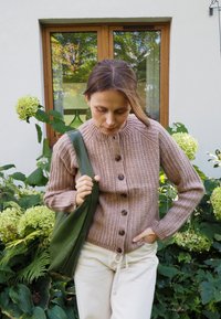 Sweter w kolorze jasnobrązowym z rypsowym nadrukiem i pięcioma guzikami; w połączeniu z zieloną skórzaną torbą i kremowymi spodniami. Lush zielone tło.