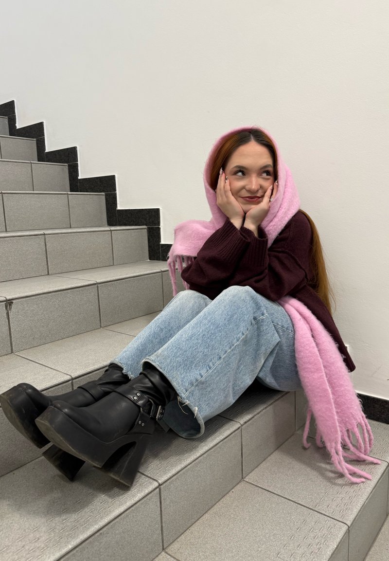 Jeune femme avec une écharpe rose assise sur des escaliers carrelés, portant un pull bordeaux, un jean bleu clair et des bottines à plateforme noires, souriant pensivement.