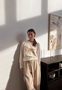 Svetlý béžový sveter s grafikou psa, kombinovaný s voľnými béžovými nohavicami. Mäkké textúry, uvoľnený strih, prírodné svetlo osvetľujúce scénu.