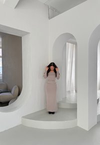 Długa, dopasowana sukienka z miękkiego beżowego materiału, z długimi rękawami i dekoltem w łódkę. Noszona z czarnymi szpilkami w minimalistycznym białym wnętrzu.