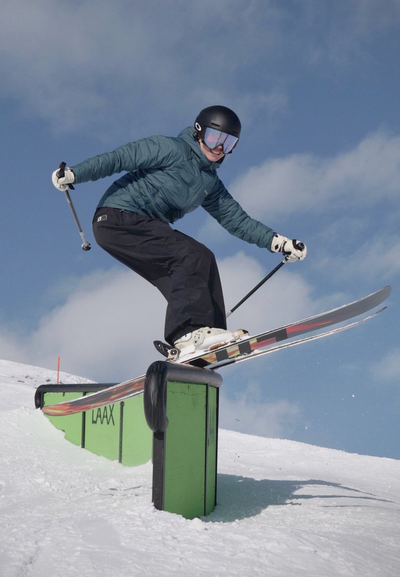 Personne portant un casque et des lunettes de protection skiant sur une structure métallique dans un parc de terrain enneigé sous un ciel nuageux.