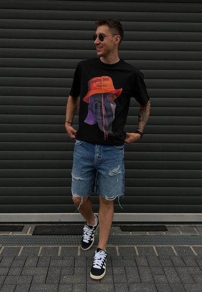 Camiseta gráfica negra con un estampado multicolor y un sombrero de cubo naranja. Combinada con pantalones cortos de mezclilla desgastados y zapatillas en blanco y negro.