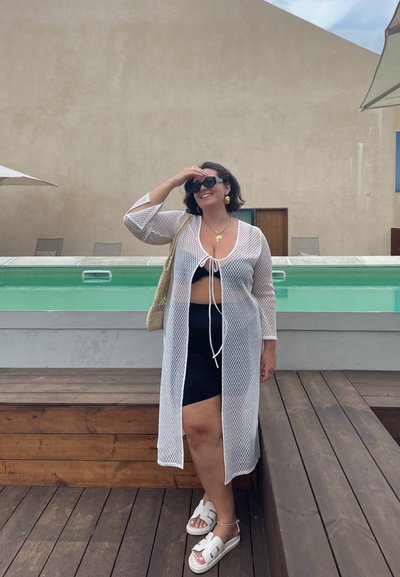 Túnica de malla blanca con mangas largas y lazo en el frente, llevada sobre un bikini negro. Sandalias blancas destacadas y un bolso de hombro de mimbre. Entorno junto a la piscina.