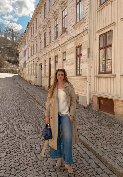 Mujer con gabardina beige y vaqueros, sosteniendo un bolso azul, de pie en una calle empedrada junto a edificios históricos color crema.