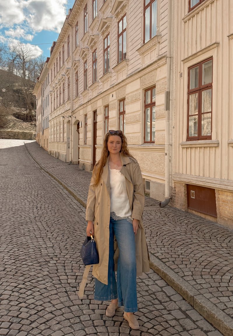 Femme en trench-coat beige et jean tenant un sac à main bleu, debout dans une rue pavée à côté de bâtiments historiques couleur crème.