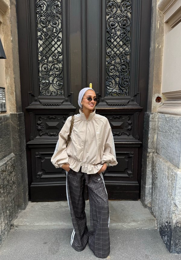 Mujer con gafas de sol, chaqueta ligera y pantalones amplios a cuadros, de pie con las manos en los bolsillos frente a una puerta de madera oscura adornada.