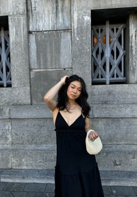 Schwarzes, ärmelloses Kleid mit rüschenbesäumtem Saum; hat ein V-Ausschnitt-Design. Hält eine kleine, geflochtene Tasche in cremefarbener Farbe. Graue Steinmauer im Hintergrund.